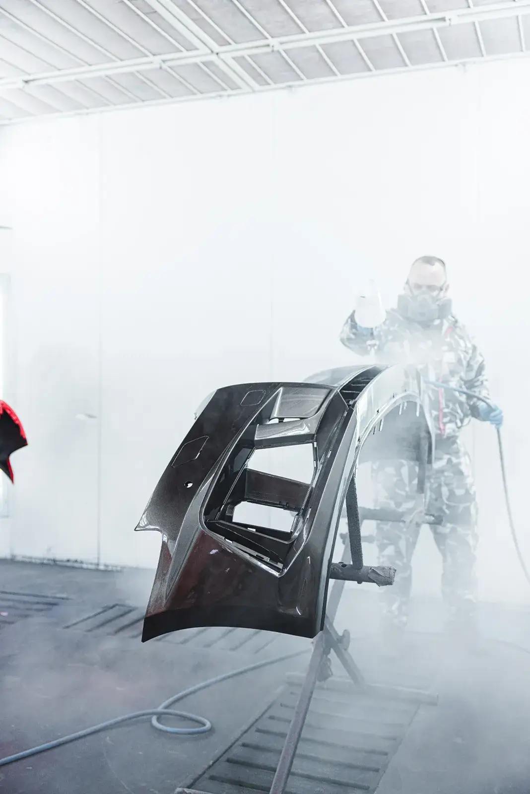 Ein Mitarbeiter in Schutzkleidung lackiert eine schwarze Stoßstange in der modernen Lackierkabine von Karosserie Blanke.